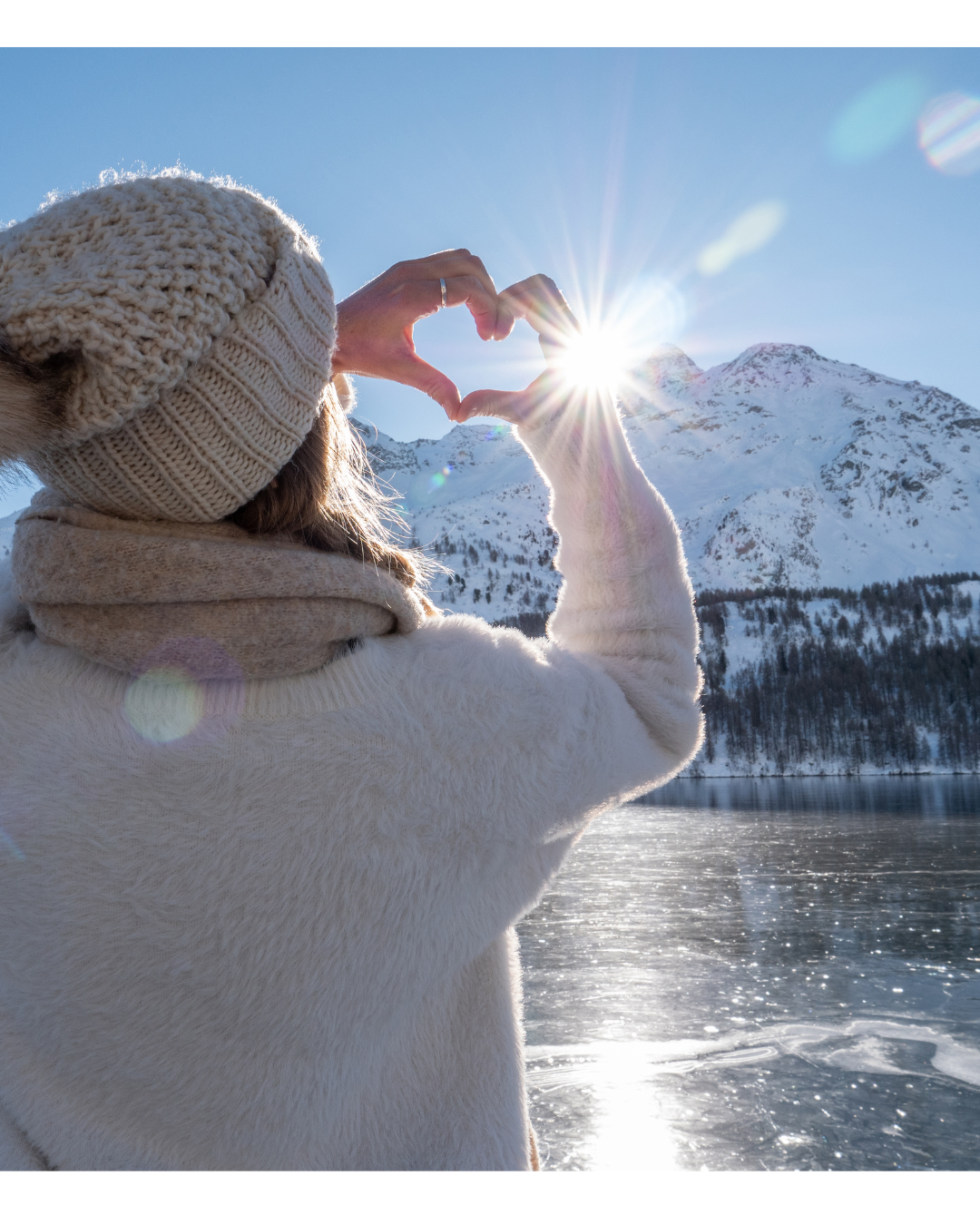 femme face à une montagne, faisant le signe d'un coeur face au soleil, habillée chaudement pour l'hiver